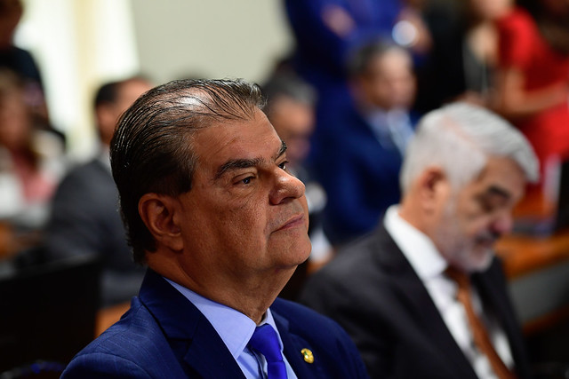 Político em evento público, discursando para uma plateia com microfone em frente.