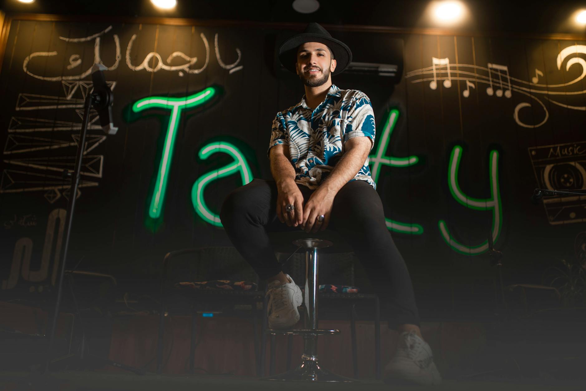 A stylish man with facial hair sits in a trendy cafe, smiling and wearing a printed shirt and hat.