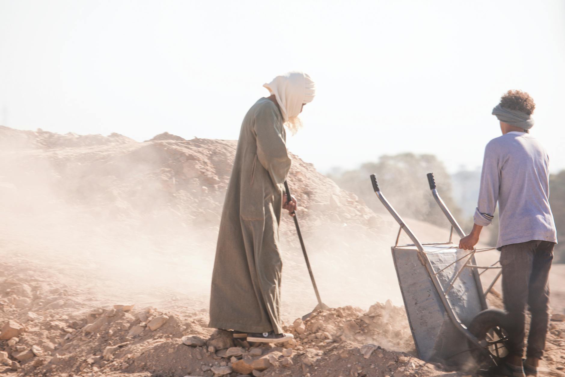 Two workers in traditional attire labor with a wheelbarrow in a dusty, arid environment.