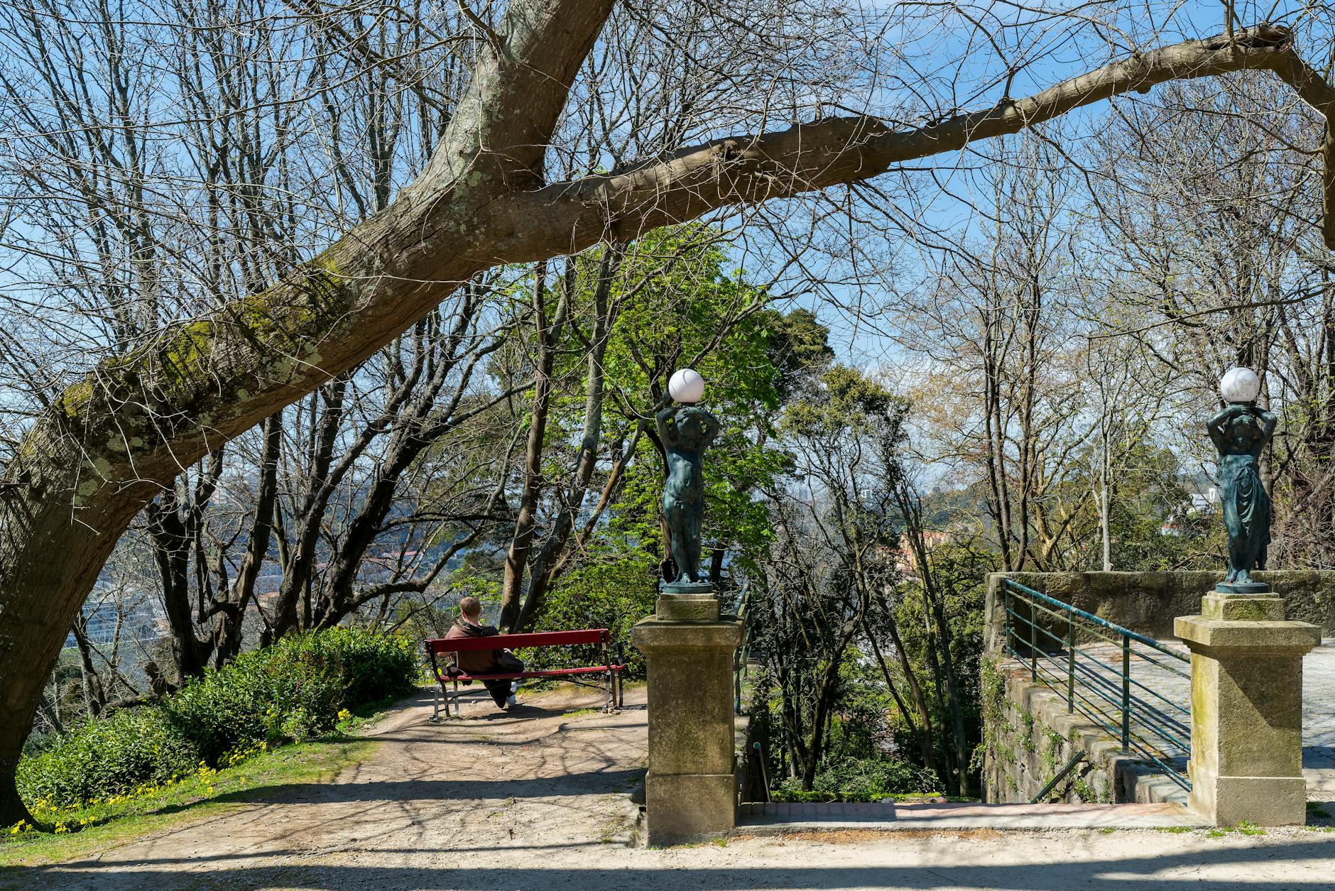 A tranquil park scene in Porto, Portugal, featuring bronze statues and a peaceful bench under trees.