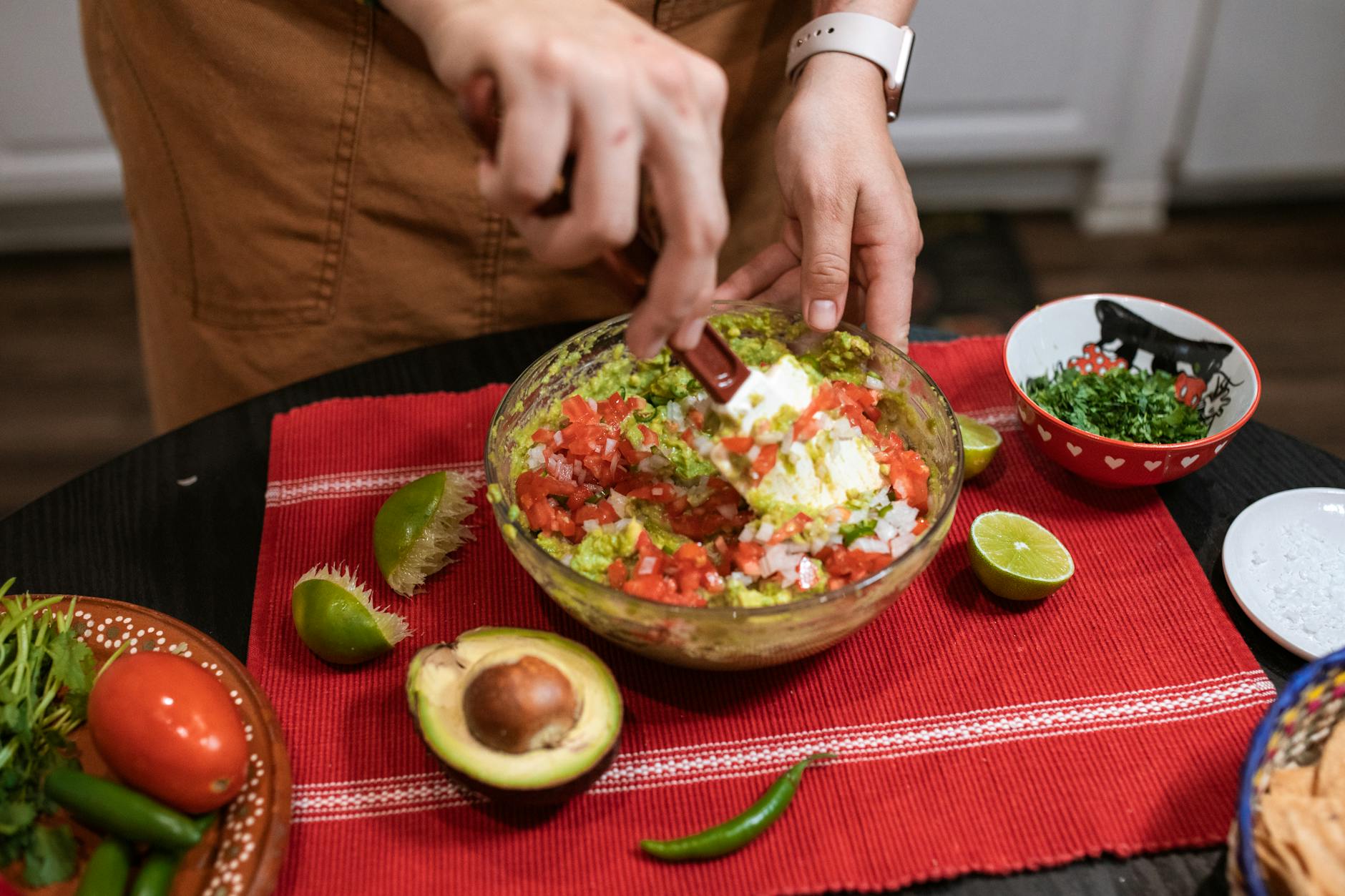 Hands preparing fresh guacamole with lime, cilantro, and tomatoes in an indoor kitchen.