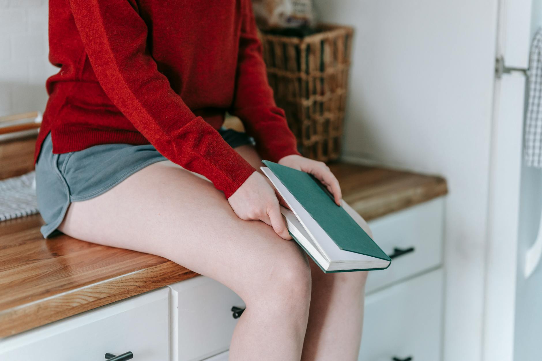A woman sits on a countertop holding a closed book in a cozy indoor setting.