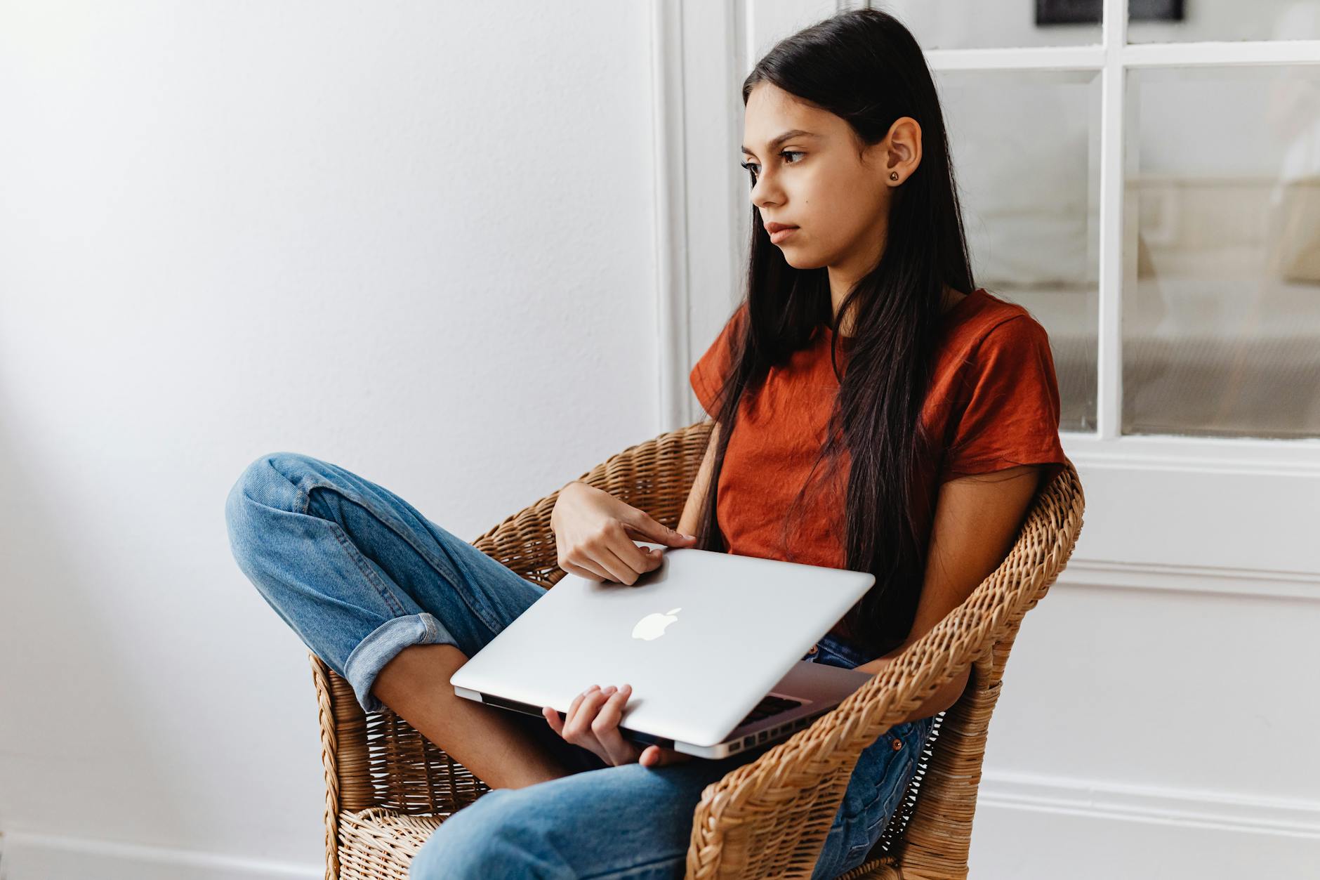 Teen girl indoors, lounging in a wicker chair, holding a laptop.