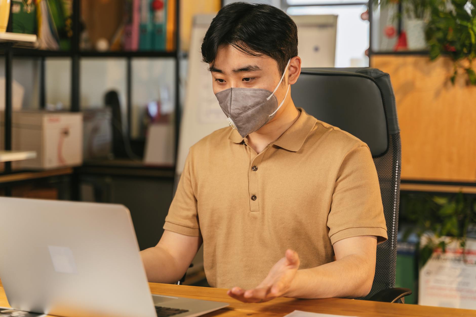 Asian man in brown shirt working at laptop, wearing a face mask in a cozy office environment.