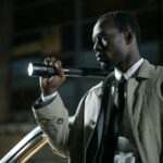African-American detective in a beige trench coat holding a flashlight outdoors at night.