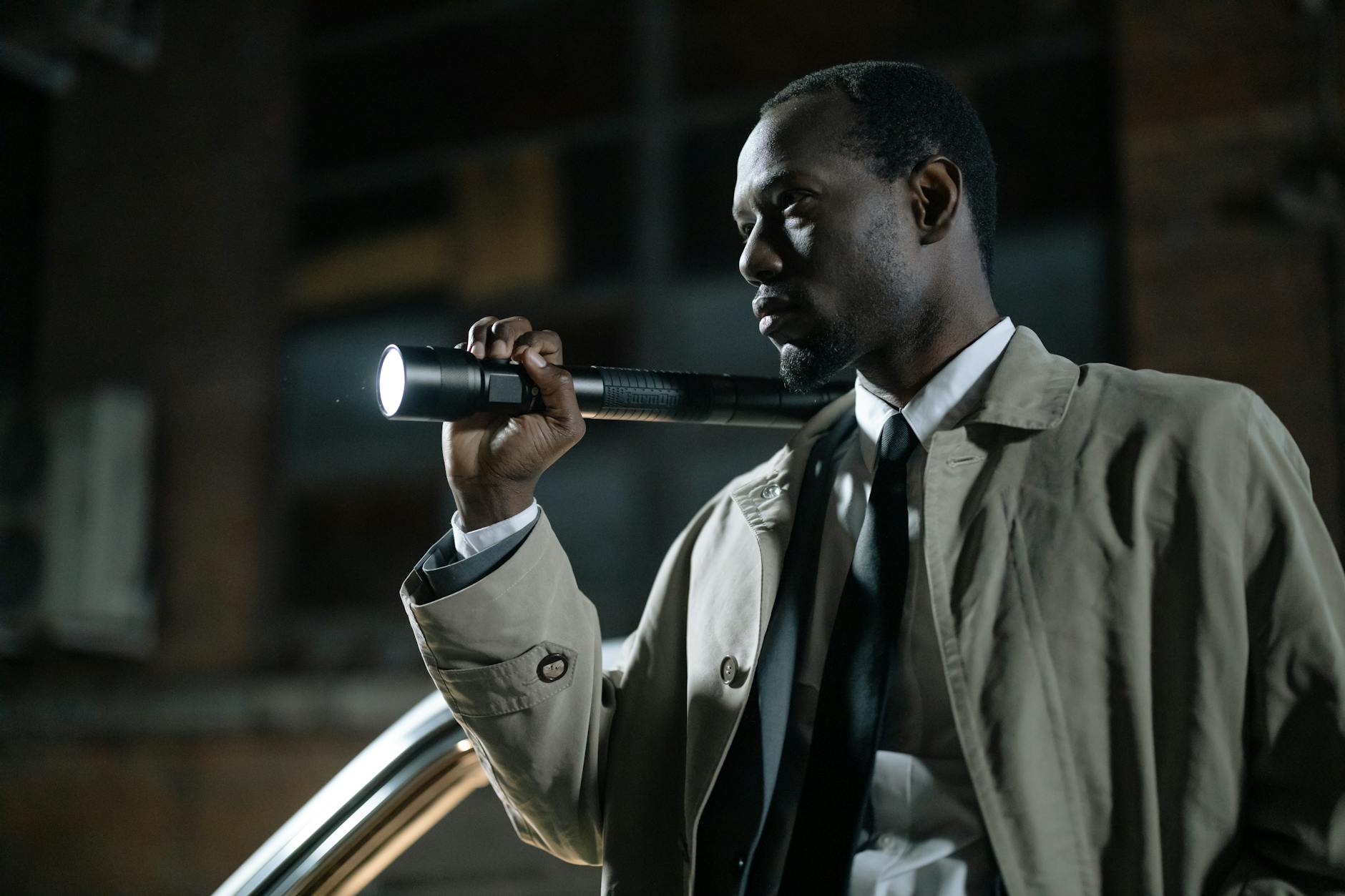 African-American detective in a beige trench coat holding a flashlight outdoors at night.