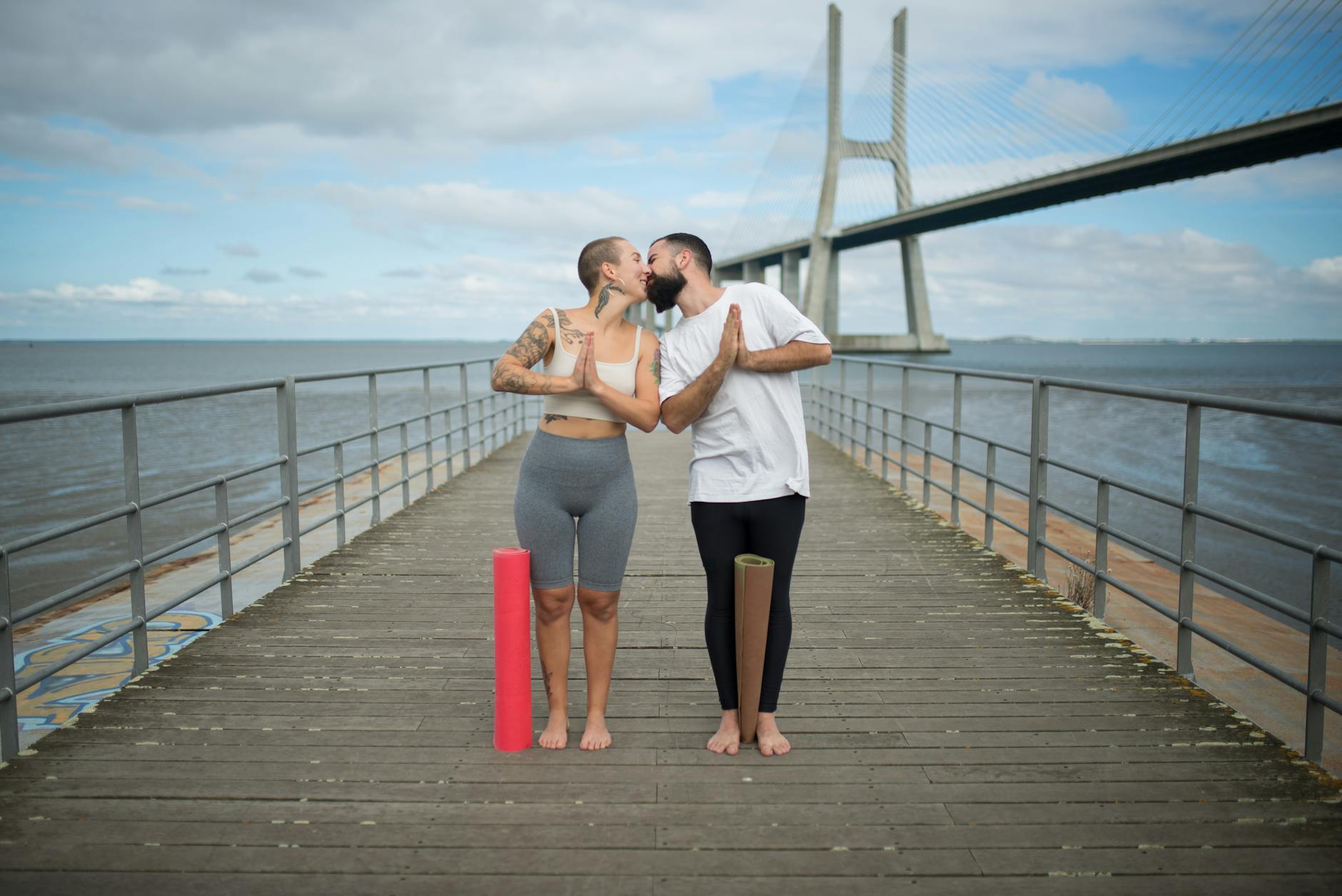 Couple sharing a romantic moment with yoga mats on a boardwalk near a suspension bridge.