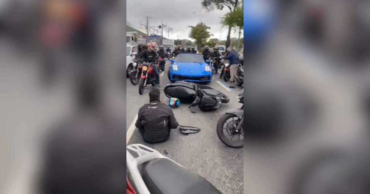 Carro esportivo de luxo parado em avenida movimentada, com pessoas e motocicletas ao redor após um incidente de trânsito.