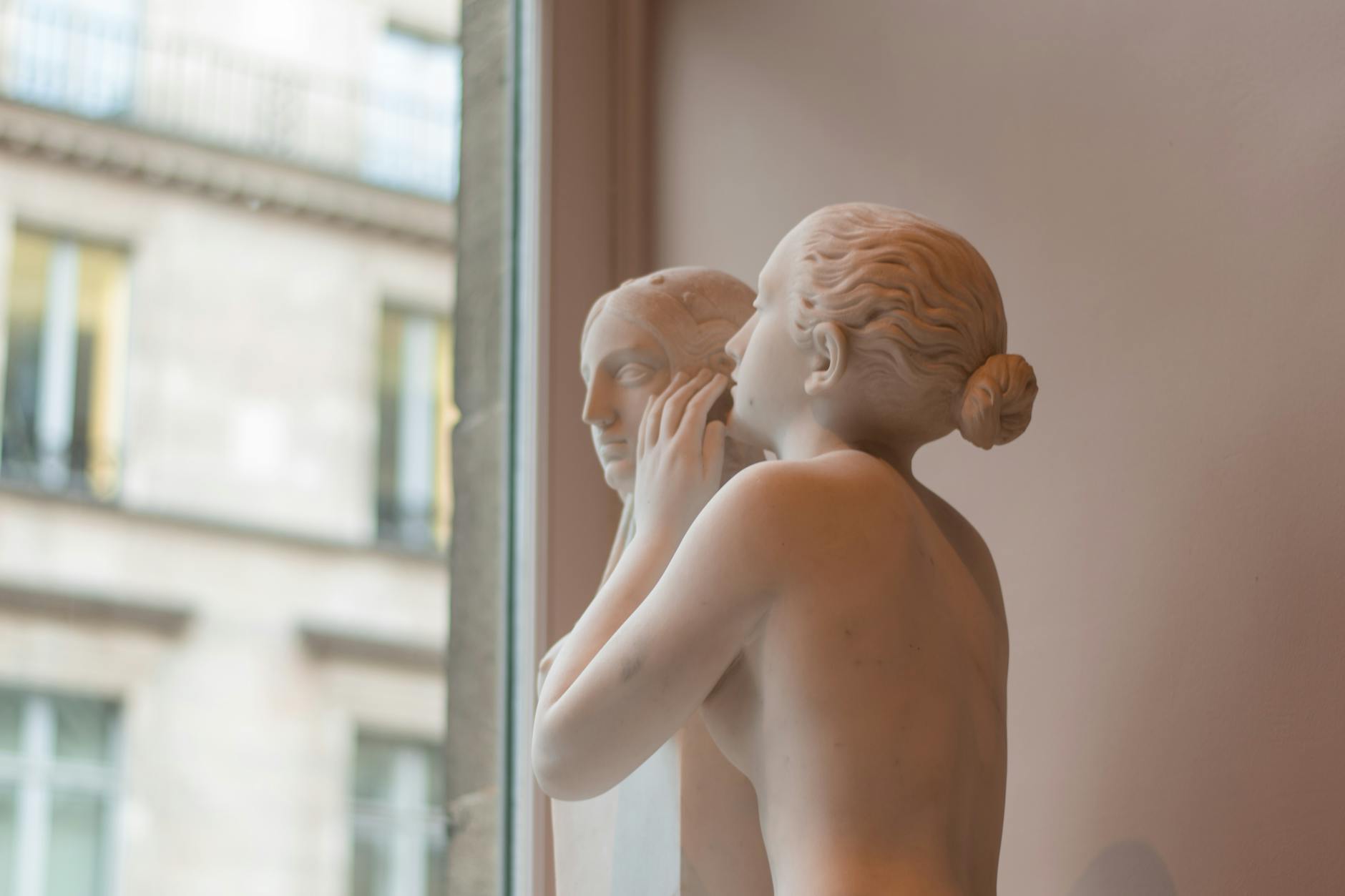 A delicate marble sculpture depicting two figures in a whispering pose by a glass window.