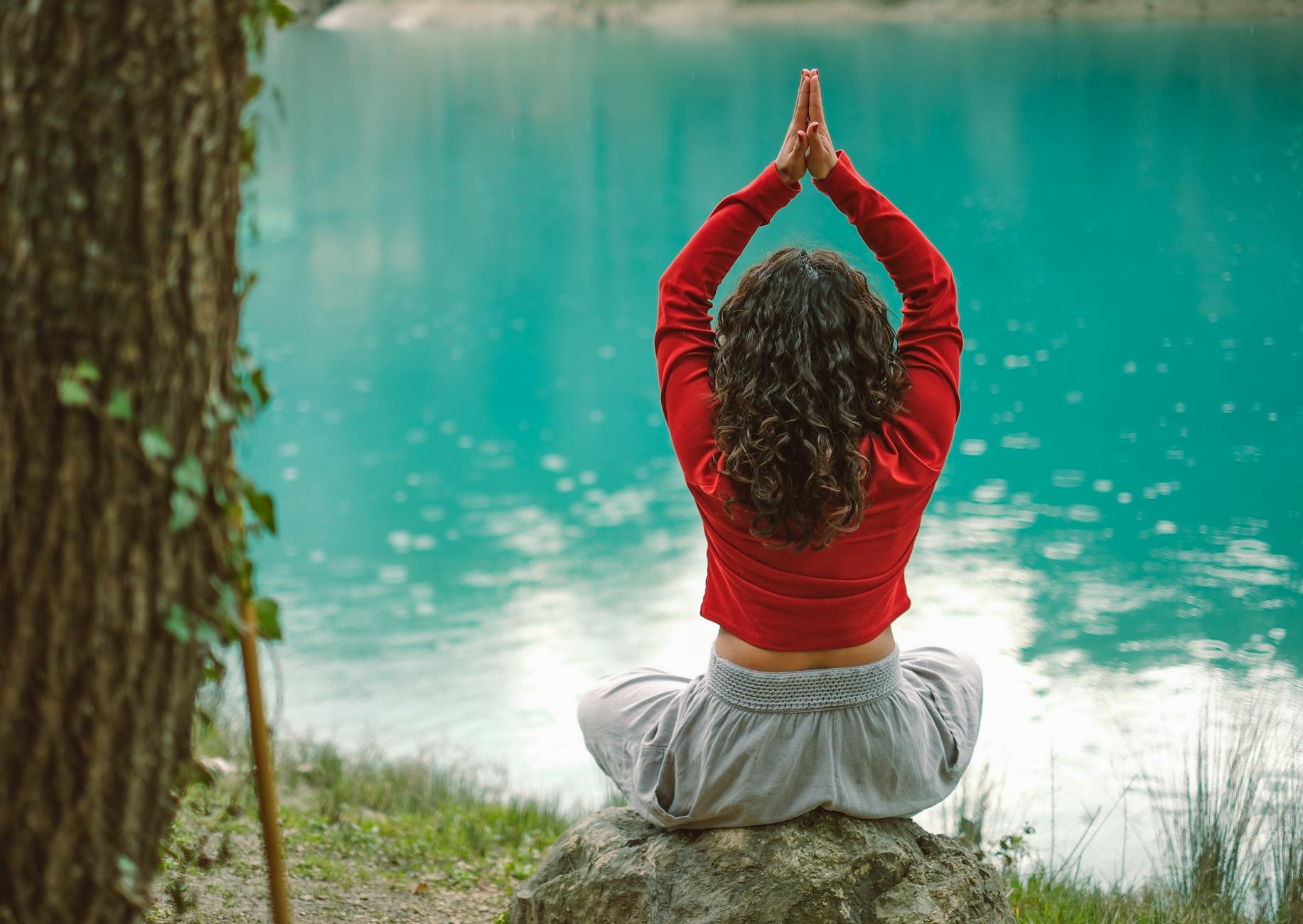 A woman meditates in nature on a rock by a tranquil turquoise lake, practicing yoga.