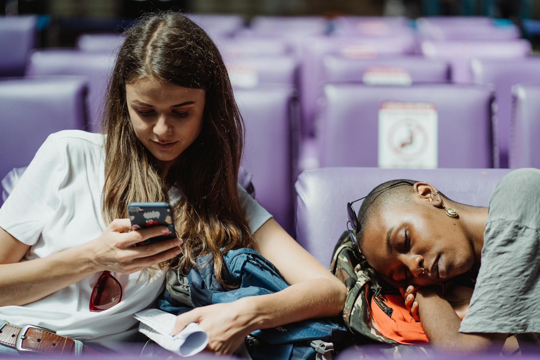 Two young women relaxing in a station. One is texting while the other is resting.