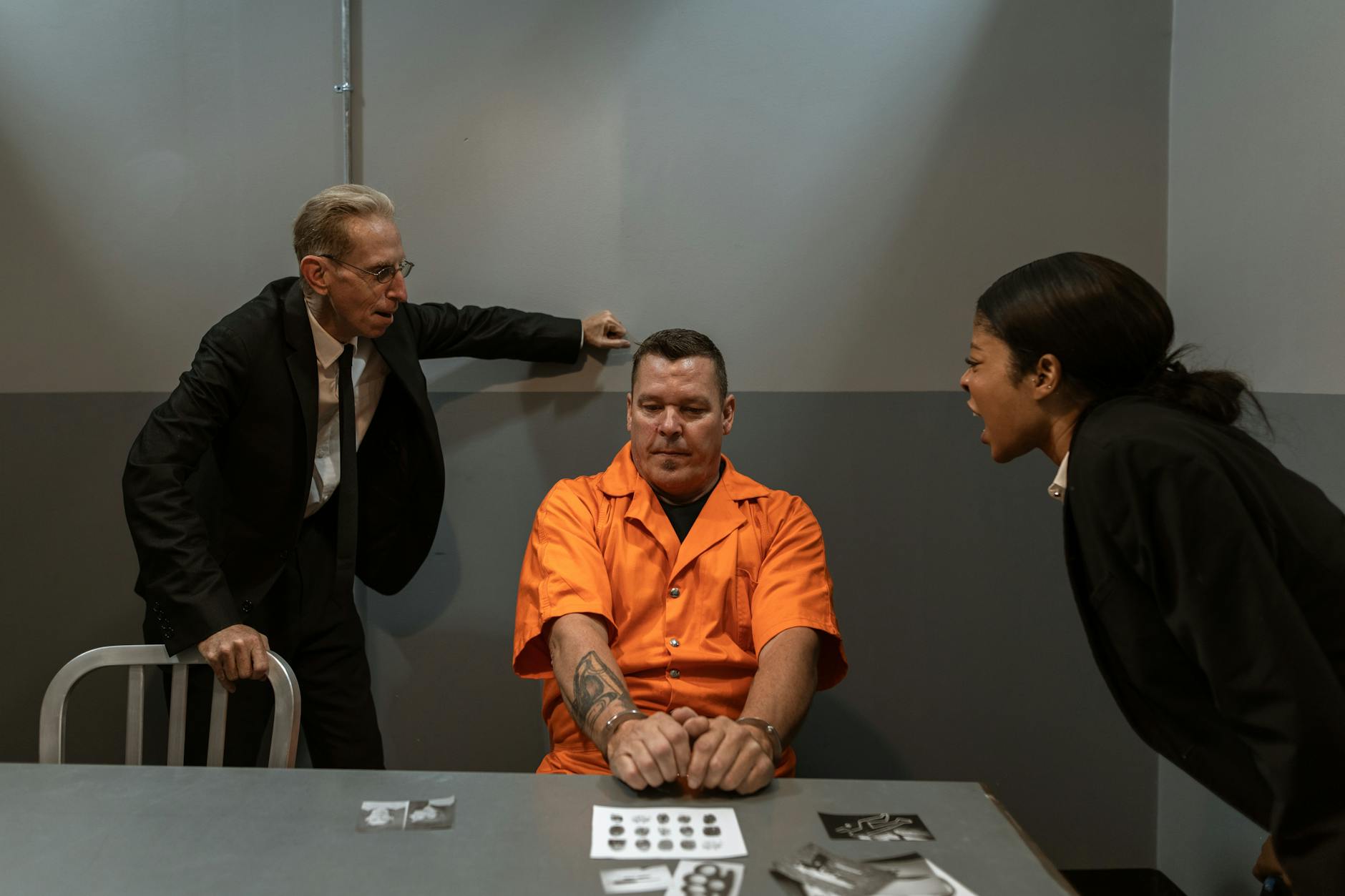 An intense interrogation with two detectives and a handcuffed prisoner in an orange jumpsuit.