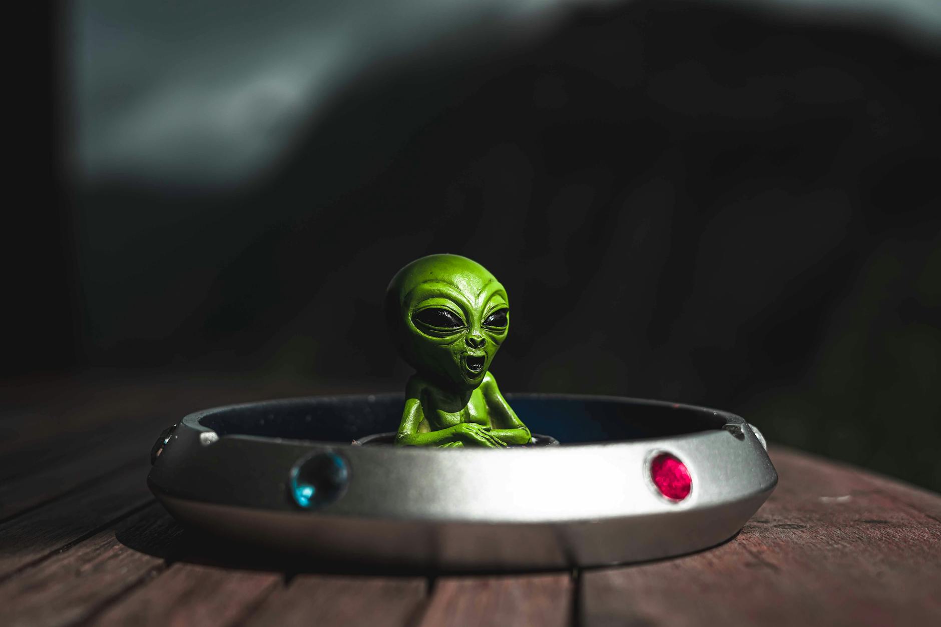 Green alien toy sitting in a spaceship-themed ashtray, outdoors with dramatic lighting.