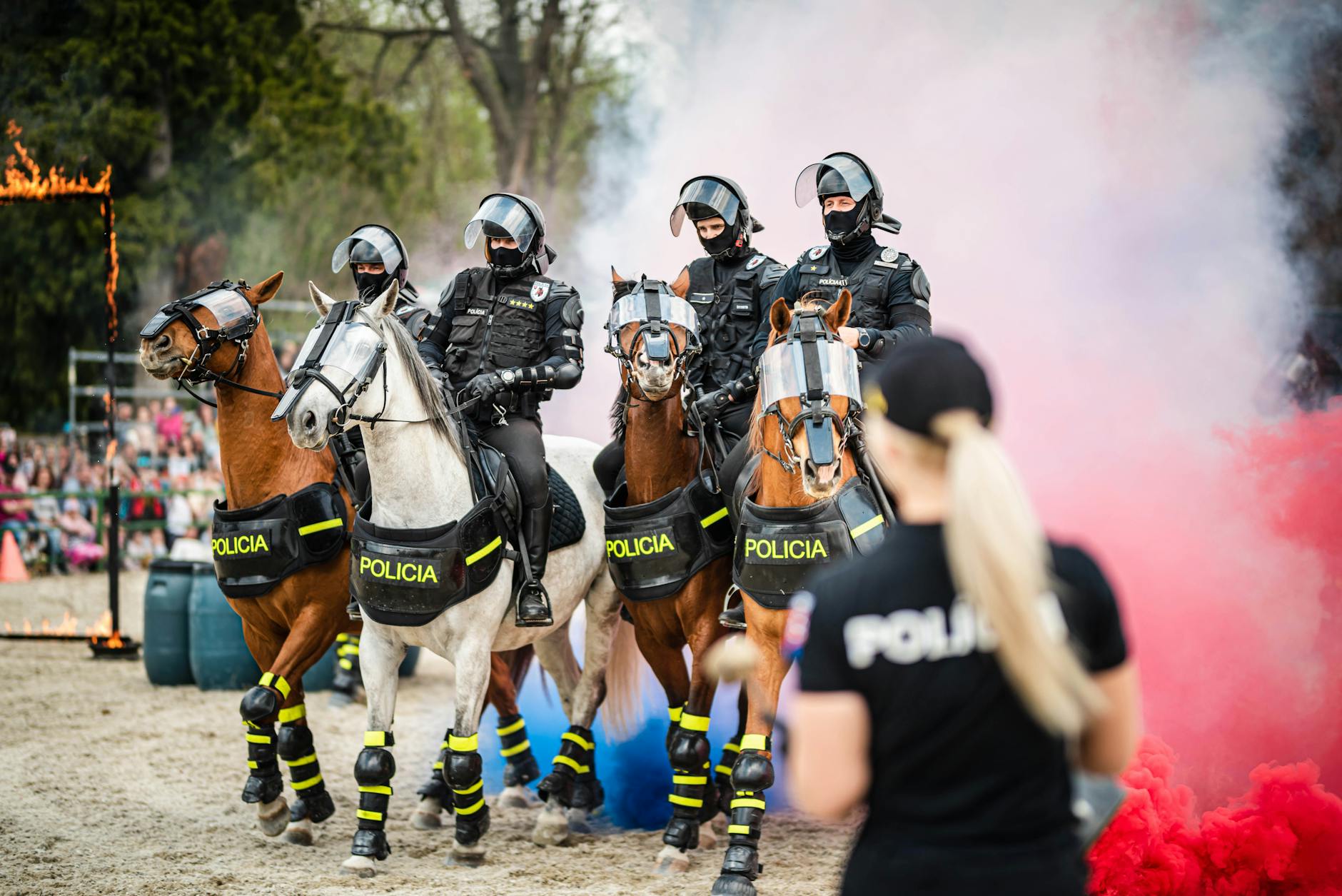 Mounted police demonstrate skills through colored smoke with crowd and flames.