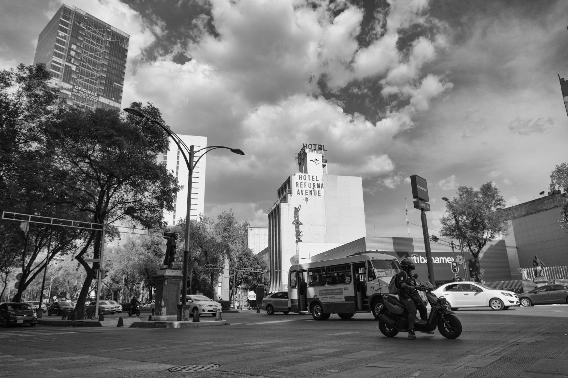 Urban street scene in Mexico City with Hotel Reforma Avenue, bus, motorbike, and trees under cloudy sky.