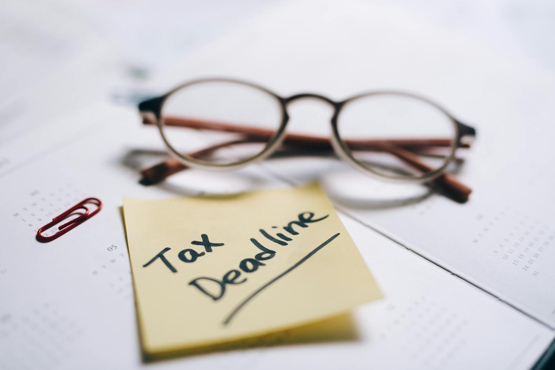 A close-up shot of a sticky note with 'Tax Deadline' written on it, alongside eyeglasses and a calendar.
